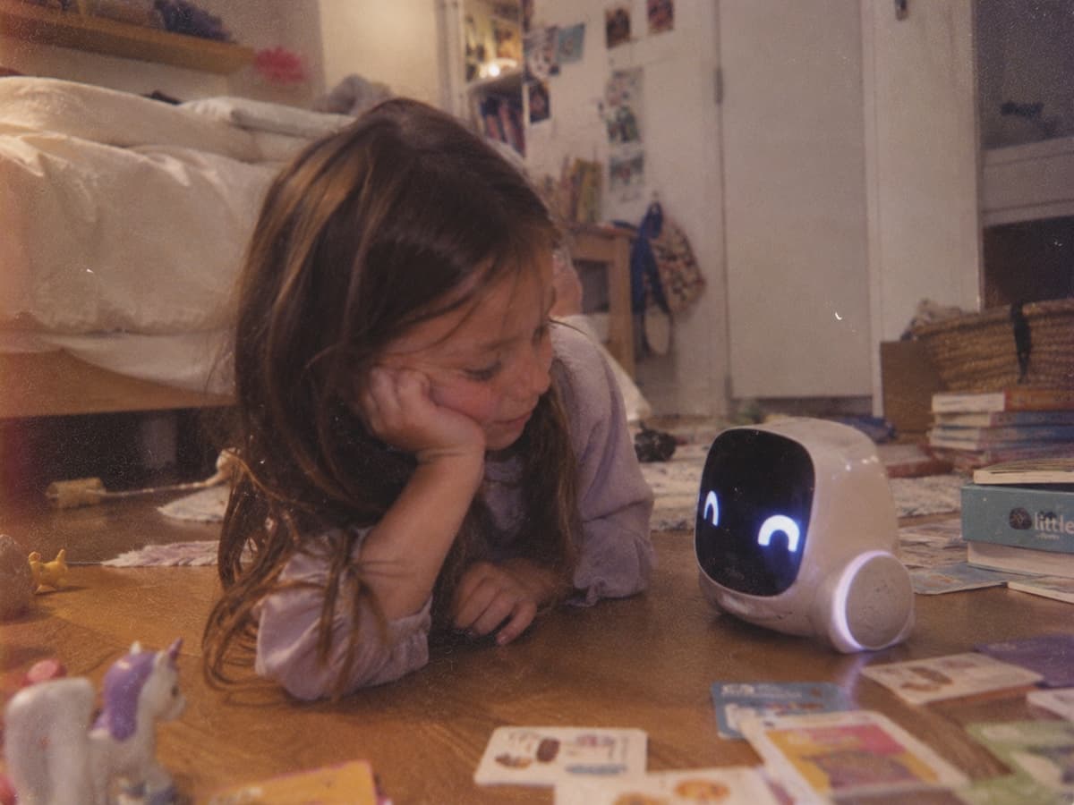 A young girl lying on her bedroom floor, chin in hand, looking at a small white robot toy with a glowing face