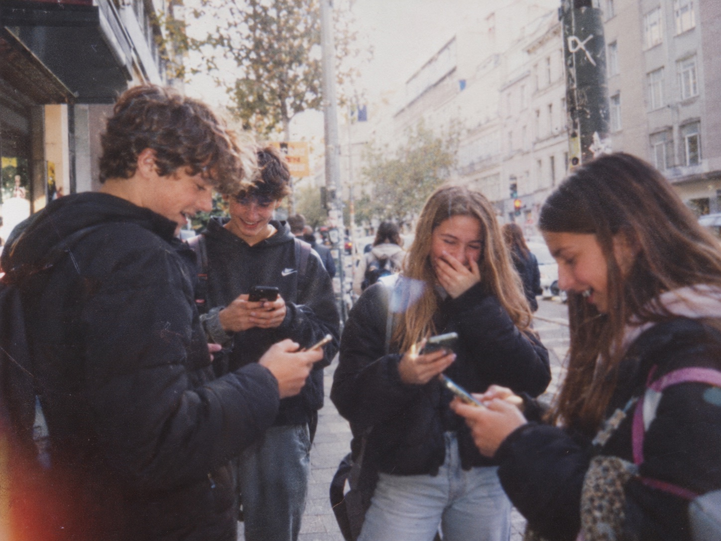 A group of teenagers on a city street, laughing as they look at their phones