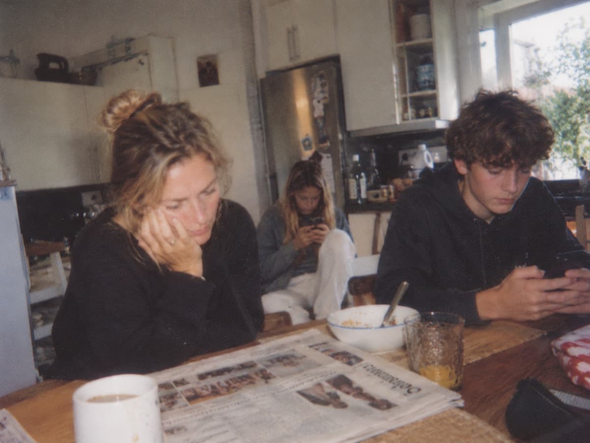 A mother resting her head on her hand at the kitchen table while her teenage son looks at his phone